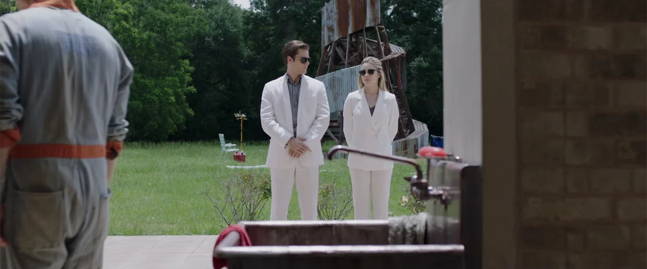 Playing God (2021) movie still: medium — A green grassy yard features a rusted metal structure and trees in the distance. Three people stand outside…
