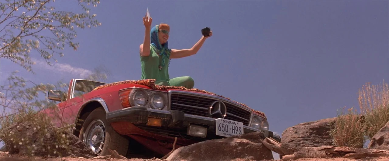 Mars Attacks! (1996) movie still: wide — A red convertible sits perched atop desert rocks under a clear blue sky. A woman in a green outfit sits on…