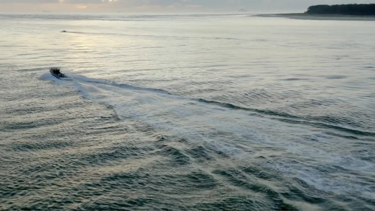 Jaws in the Shallows (2023) movie still: wide — A vast expanse of water under an overcast sky at dusk with distant land. A small motorboat travels…