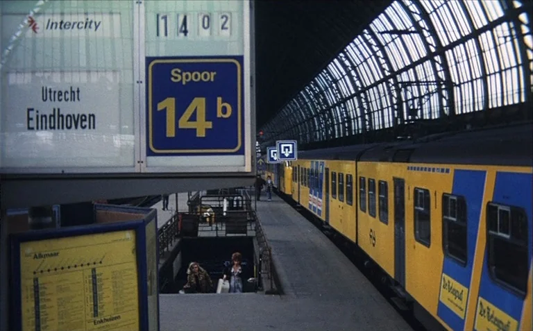 Business is Business (1971) movie still: wide — A spacious railway station platform features a large yellow and blue Dutch train parked. An…