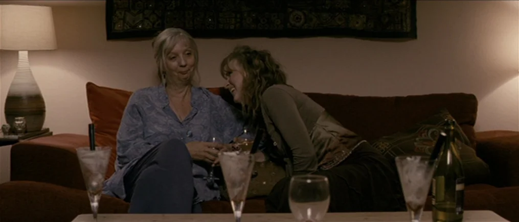 Another Year (2010) movie still: medium — A dimly lit room features a rust-colored couch adorned with a patterned textile wall hanging. Two women…