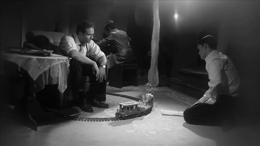 Keyhole (2011) movie still: wide — A dark, sparsely furnished room with a small electric train circling on the carpet. Two men sit on the floor and…
