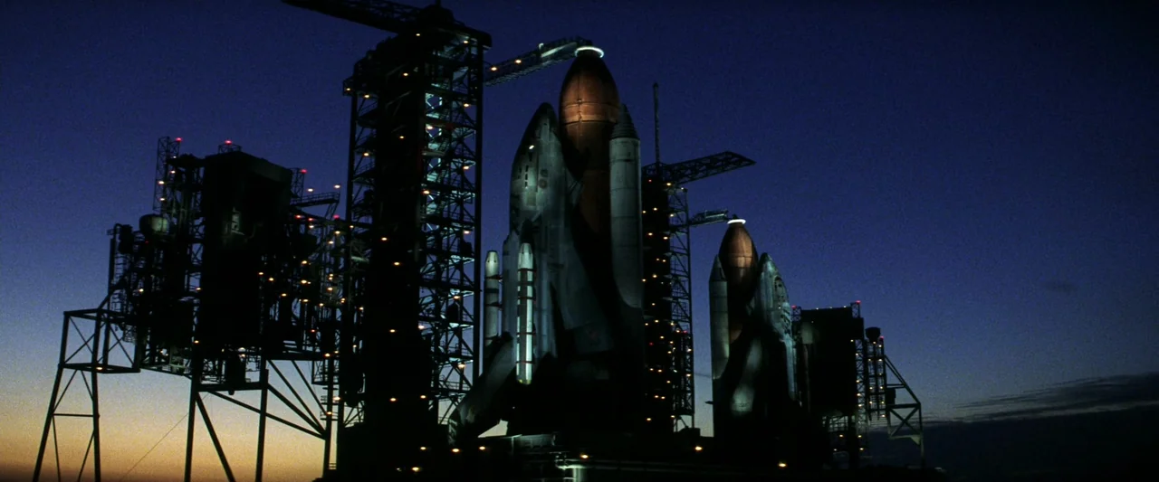 Armageddon (1998) movie still: establishing — Two space shuttles are mounted on towering metal gantries at a dark launch facility. The horizon glows…