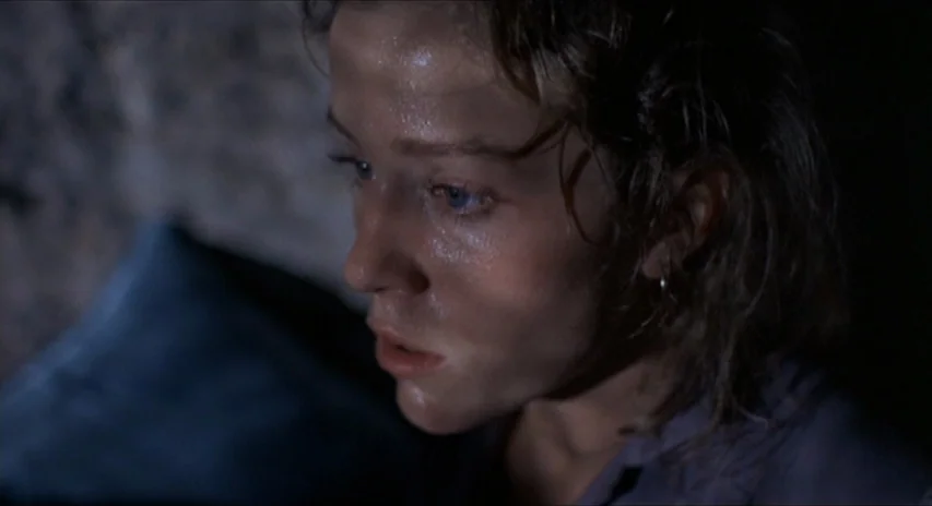 Blood Simple (1984) movie still: closeup — A dark, dimly lit environment with a rough textured stone wall in the background. A young woman with a…