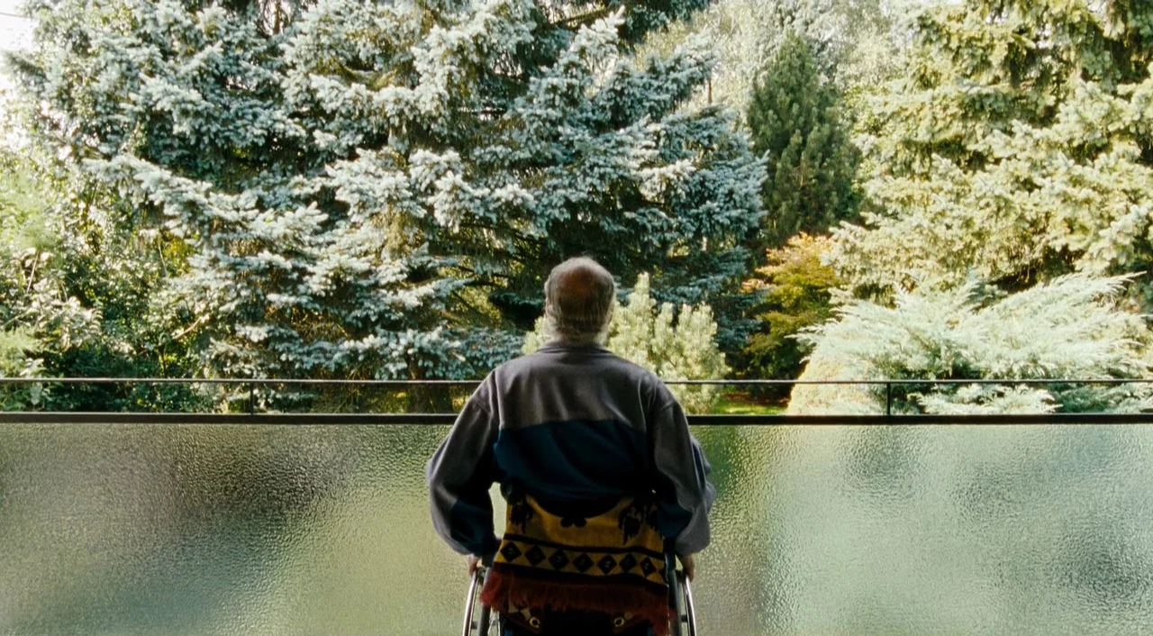 Paradise: Faith (2012) movie still: medium — A man sits on an outdoor balcony enclosed by a textured glass railing. He faces a dense screen of tall…