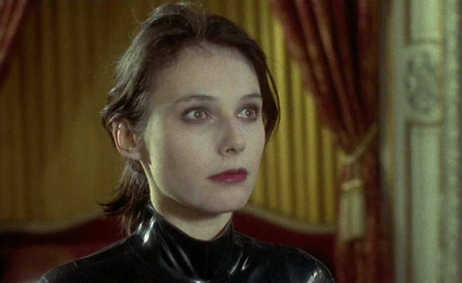 Irma Vep (1996) movie still: closeup — A woman stands in front of gold-colored fabric curtains inside a room. She looks off-camera while wearing a…