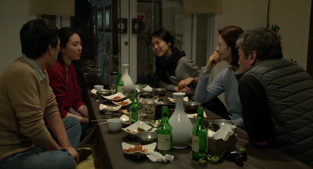 On The Beach At Night Alone (2017) movie still: medium — Five people sit around a table laden with food and green soju bottles. They are engaged in…