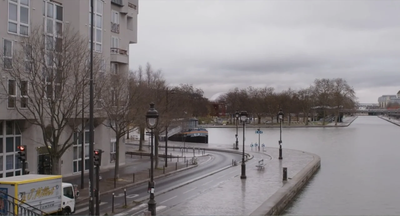 A Dramatic Film (2019) movie still: establishing — A stone embankment borders a calm canal beside a tall apartment building on a day. The curved…