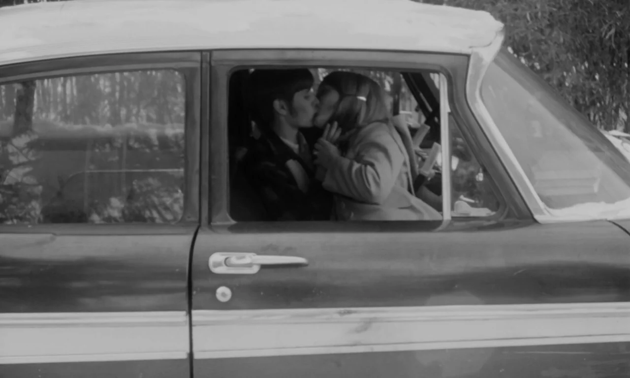 Multiple Maniacs (1970) movie still: medium — A vintage 1960s car is parked near trees on a bright day. A young man and woman are sitting in the…
