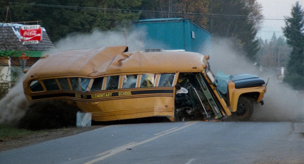 Out of the Blue (1980) movie still: wide — A damaged school bus sits wrecked on a paved rural road with dust clouds rising. A blue truck trailer is…