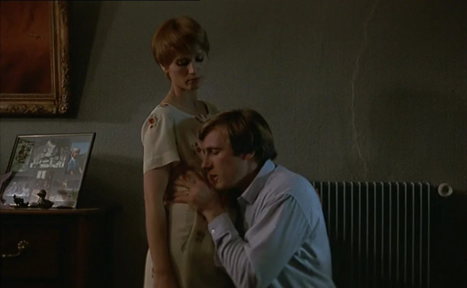 The Woman Next Door (1981) movie still: medium — A dimly lit room with textured dark walls features a side table with photo frames. A man kneels in…