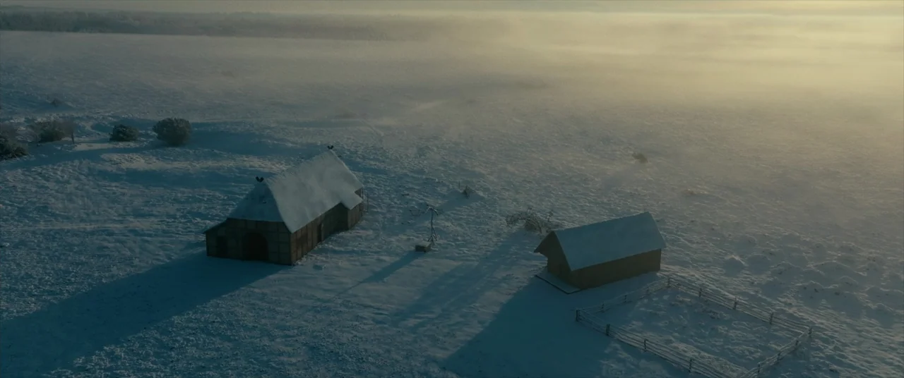The Promised Land (2023) movie still: establishing — Two wooden structures stand in an expansive, flat, snow-covered landscape under a morning fog.…
