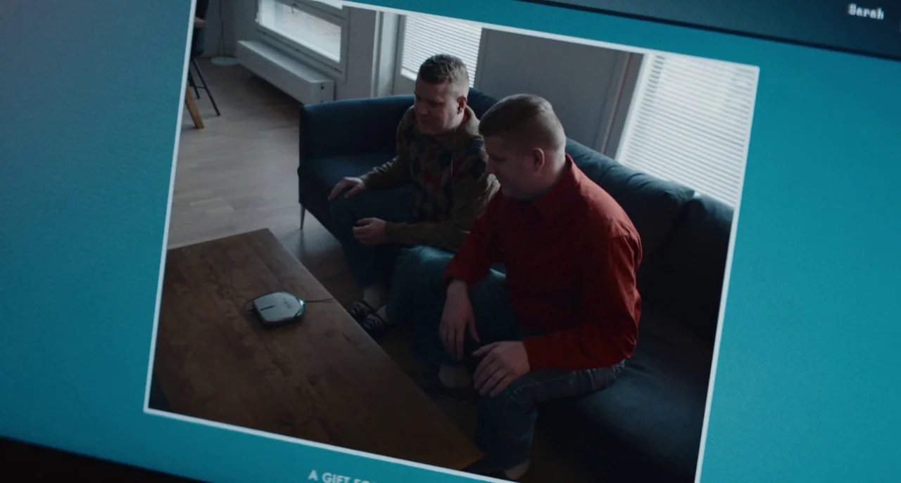 Dual (2022) movie still: medium — Two men sit on a dark sofa in a modern room with wooden flooring. A small electronic device rests on a wooden…