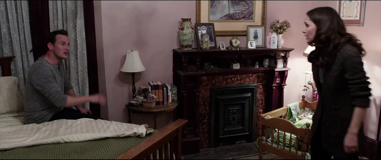 Insidious: Chapter 2 (2013) movie still: medium — A dimly lit room with pink walls, a dark wooden fireplace, and vintage decor. A man sits on a bed…