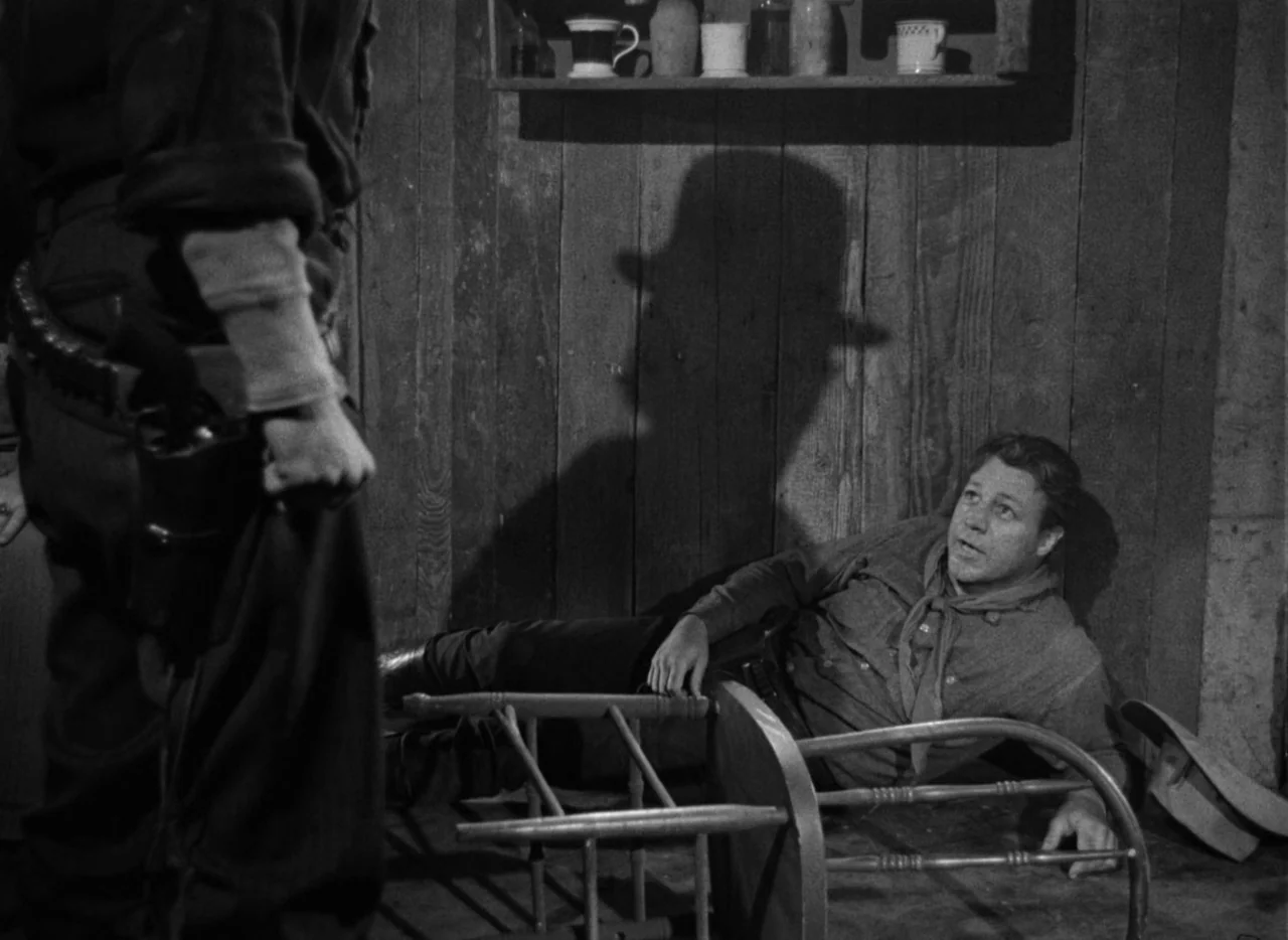 Red River (1948) movie still: medium — A wooden interior with vertical plank walls features a shelf with various jars. A man sits on the floor near…