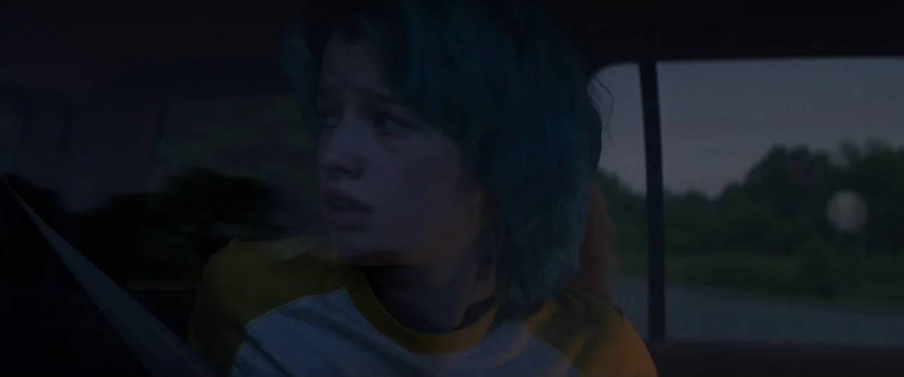 Black Widow (2021) movie still: closeup — The interior of a car at dusk with a young woman in the backseat. She gazes out of the side window while…