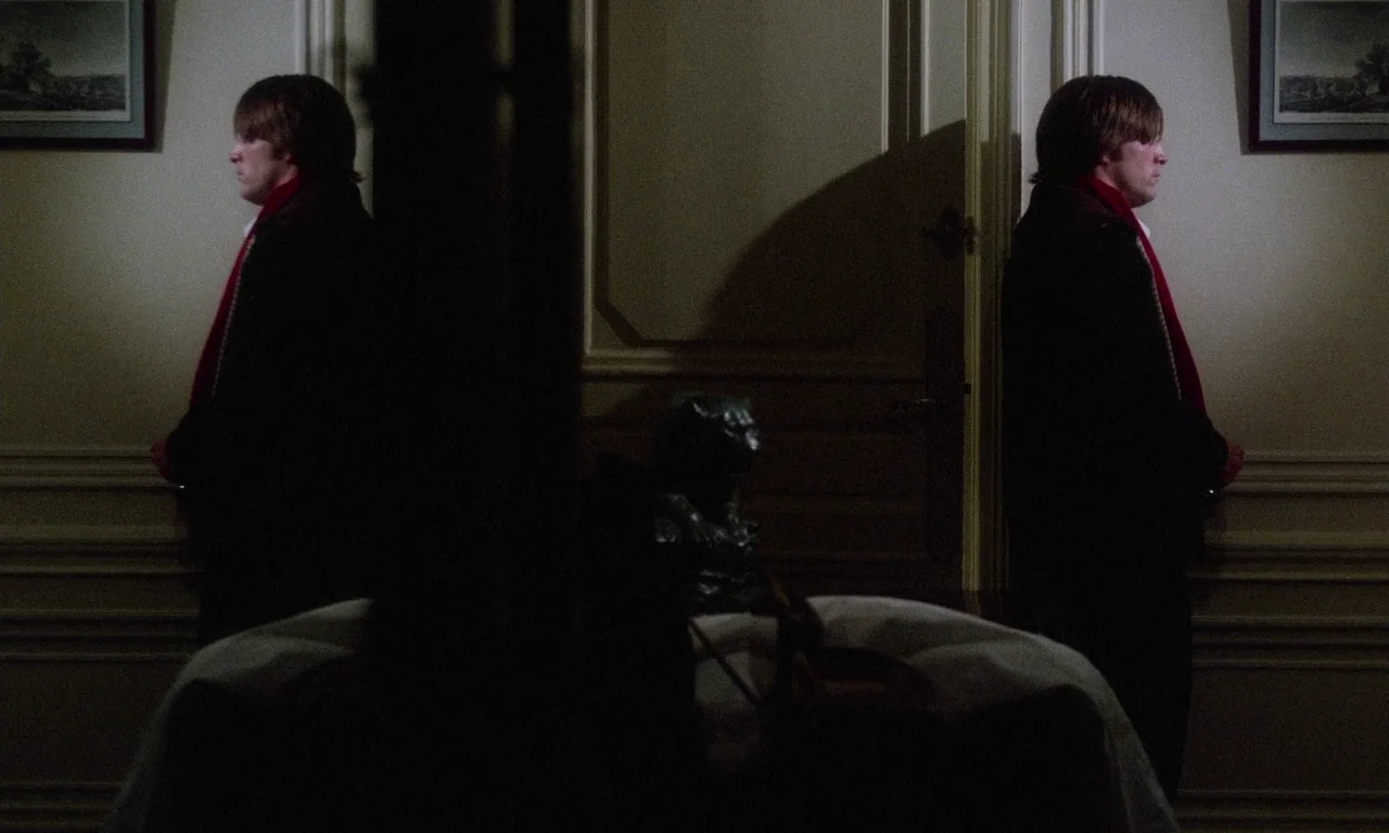 The Margin (1976) movie still: medium — A symmetric interior hallway features beige walls adorned with framed landscape paintings and molding. Two…