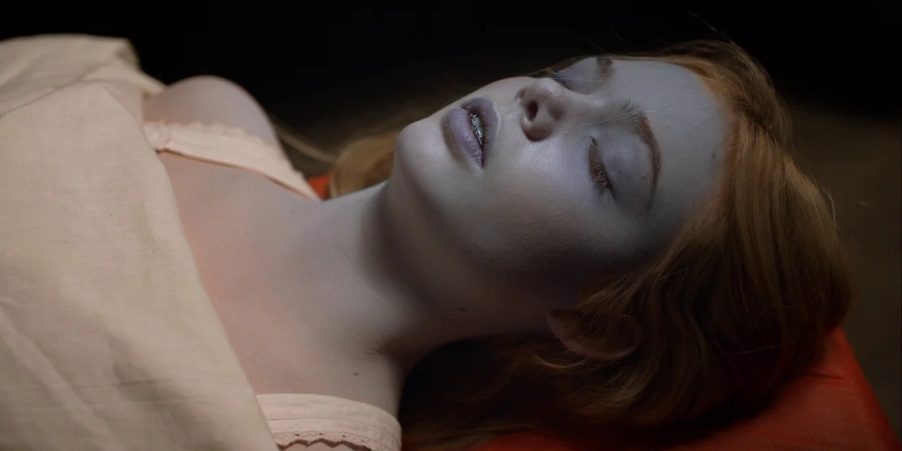 Twixt (2011) movie still: closeup — A young person with pale skin lies on an orange surface under a sheet. The person has closed eyes and visible…