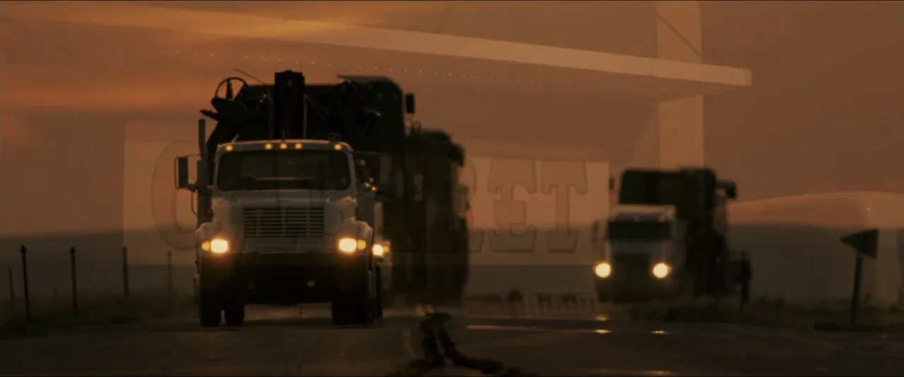 Into the Wild (2007) movie still: wide — A long asphalt road stretches across an open plain under a hazy golden sky. Two large semi-trucks travel…
