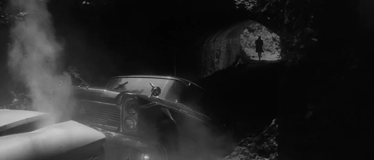 Branded To Kill (1967) movie still: wide — A dark underpass frame shows a smoking wrecked vintage car in the foreground shadows. A man runs through…