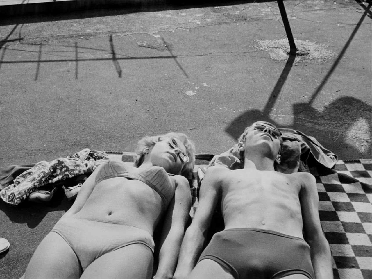 The Sun In A Net (1963) movie still: medium — A flat concrete rooftop surface is marked by long, sharp shadows from overhead structures. A young man…