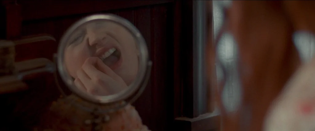 In A Valley of Violence (2016) movie still: closeup — A dimly lit room features a small vanity mirror reflecting a woman's face. The woman observes…