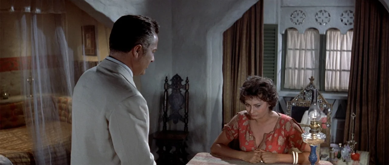 Legend of the Lost (1957) movie still: medium — A rustic interior with thick white plaster walls and dark wood window coverings. A man in a light…