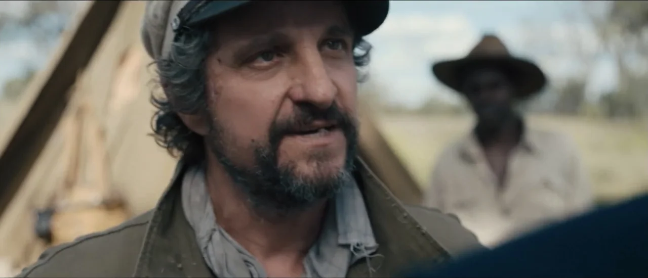 Fear Below (2025) movie still: closeup — A rural campsite features a canvas tent structure under an overcast sky. A middle-aged man with a beard…