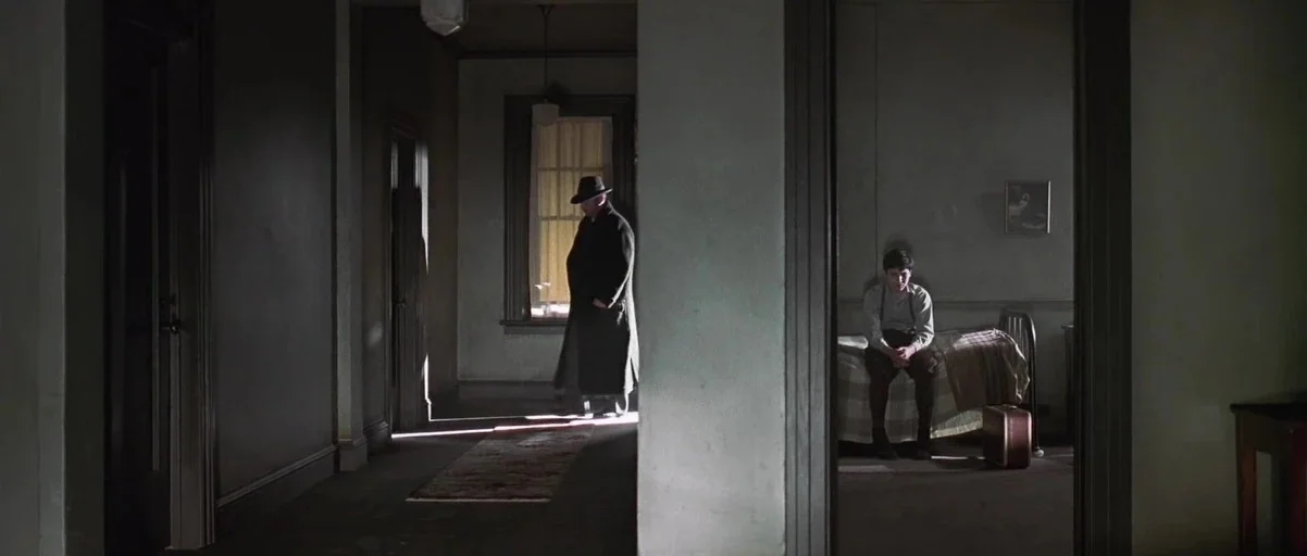 Road To Perdition (2002) movie still: wide — A dimly lit interior corridor features a man in a coat standing by a window. In the adjacent room, a…