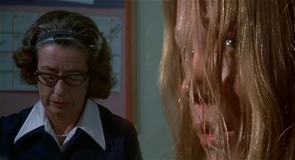Carrie (1976) movie still: medium — An interior office space with a wall calendar visible behind a middle-aged woman. The woman sits opposite a…