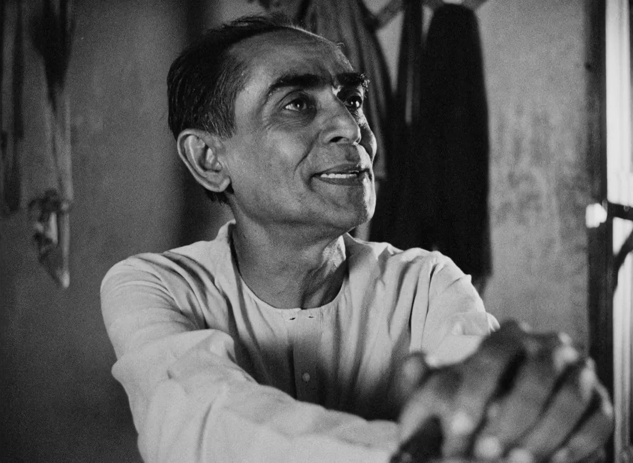 Apur Sansar (1959) movie still: medium — An interior space features simple walls with clothing hanging in the background blurred. A man sits while…