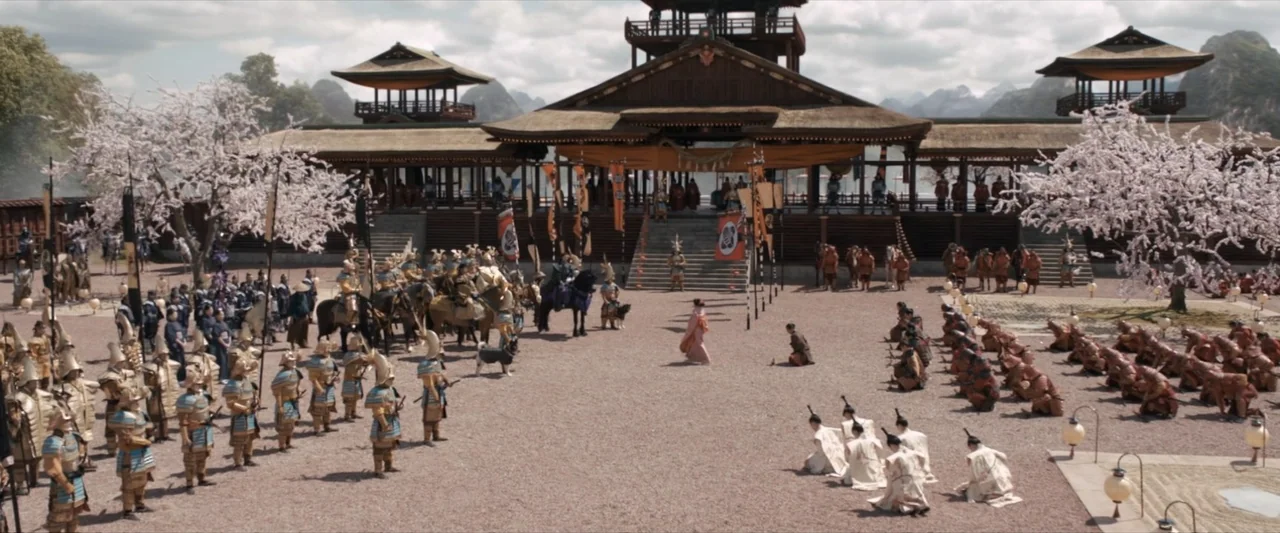 47 Ronin (2013) movie still: wide — A spacious courtyard surrounds a large wooden temple building with tiered roofs and cherry blossoms. Soldiers in…