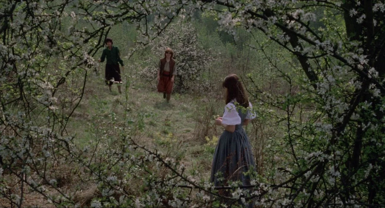 The Blood on Satan’s Claw (1971) movie still: wide — A grassy meadow surrounded by blooming white flowered bushes in early spring. Three young…
