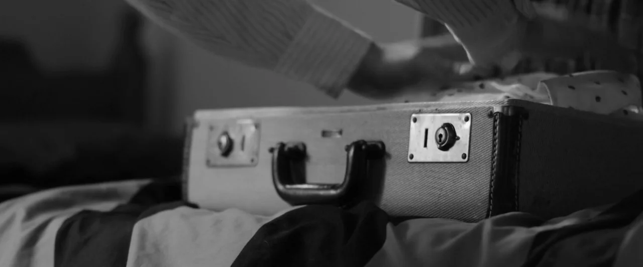 Weirdos (2016) movie still: closeup — A close-up shot focuses on a vintage suitcase resting on a rumpled bed sheet. An individual's hands reach into…