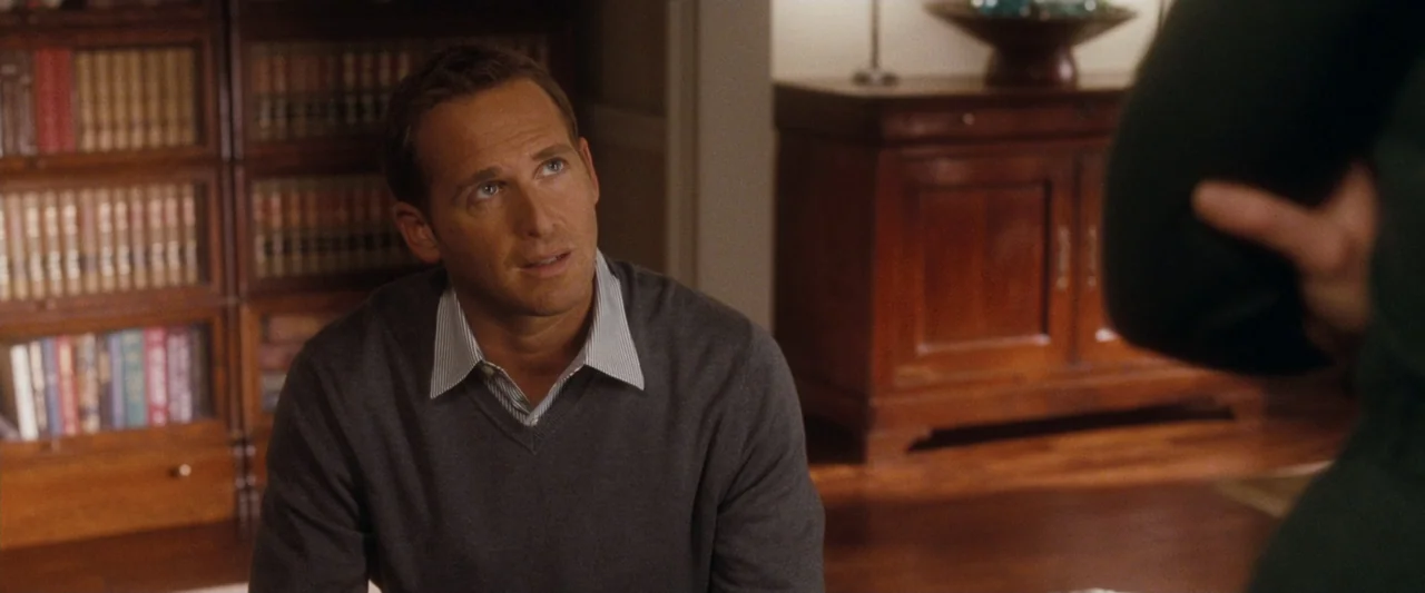 Life As We Know It (2010) movie still: medium — A dimly lit home office features a wooden bookcase filled with books in the background. A man in a…
