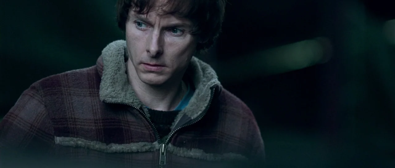 Isolation (2005) movie still: medium — A dark outdoor setting, likely a forest at night, obscured by shadows. A man stares intensely off-screen,…