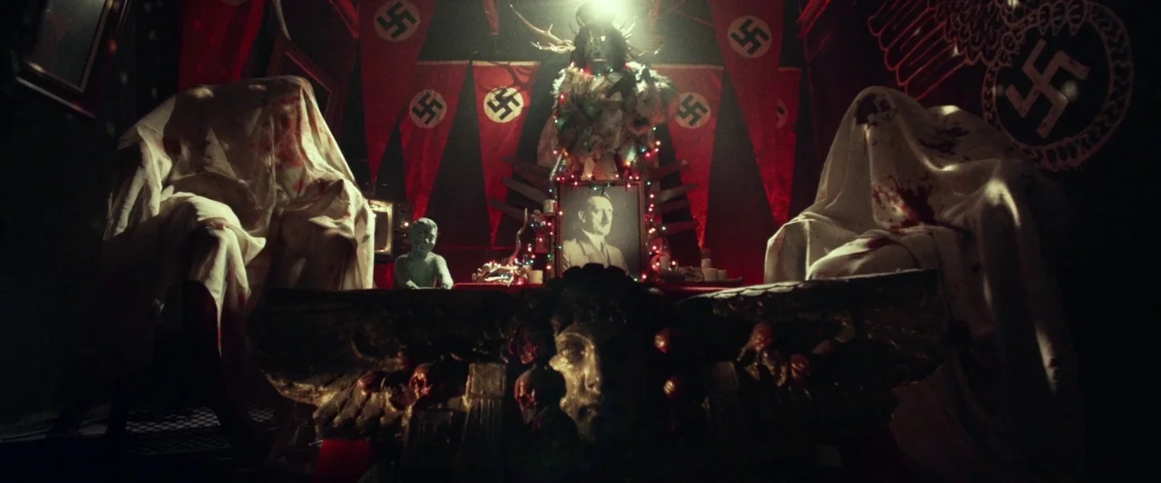 31 (2016) movie still: wide — A dark, cramped chamber decorated with red banners featuring Nazi symbols and central altar. Two figures draped in…