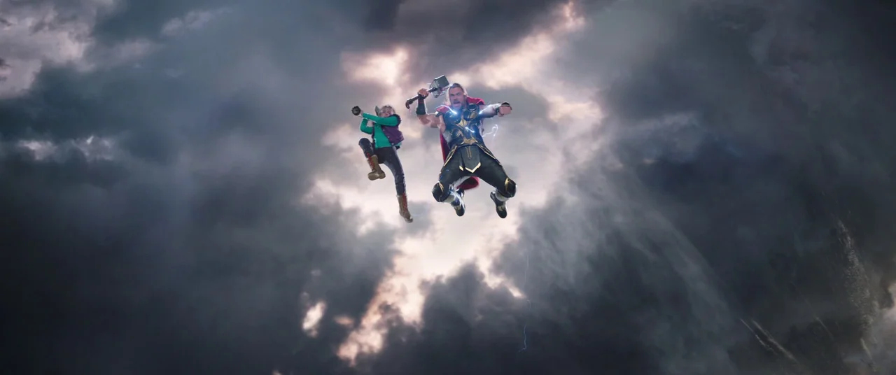 Thor: Love and Thunder (2022) movie still: wide — Two individuals fly through a heavy, dark cloud-filled sky. They are suspended in mid-air amidst a…
