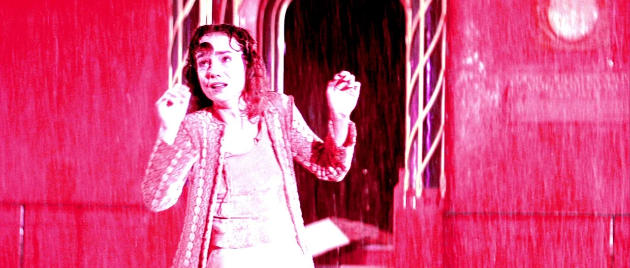Suspiria (1977) movie still: medium — A woman stands under heavy rainfall in front of a dark building doorway. She looks upward with an expression…