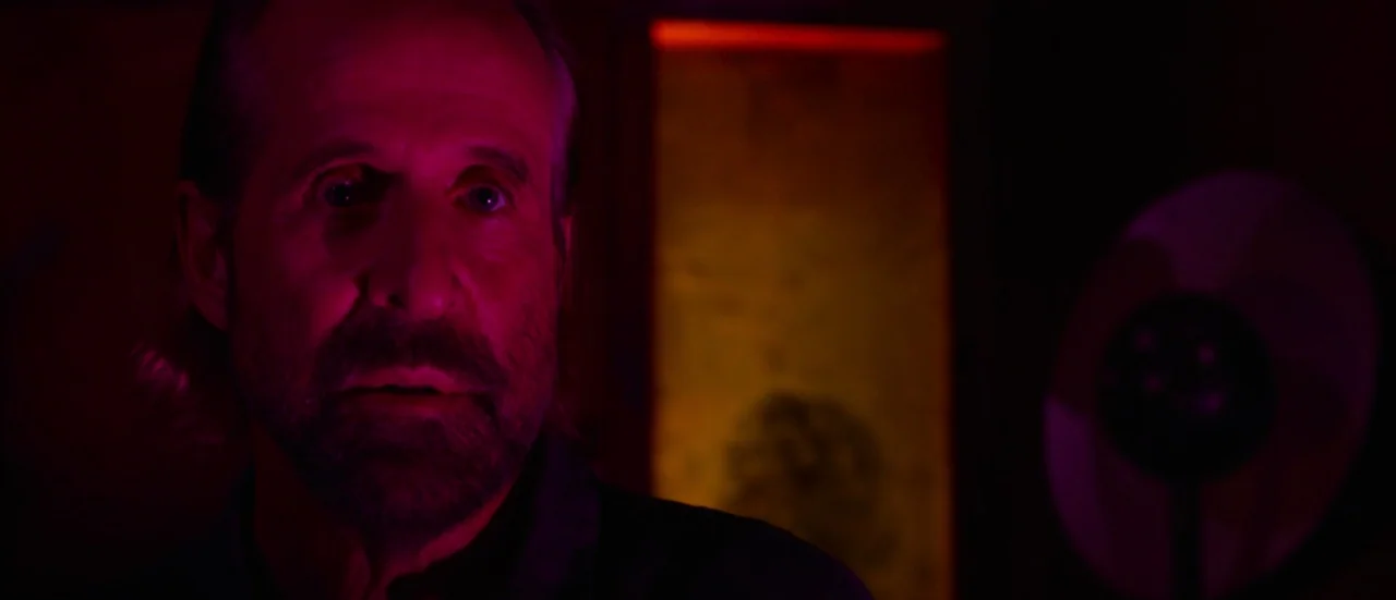 Rupture (2016) movie still: closeup — An older man is positioned in a dimly lit space with saturated red lighting. He is looking toward the side of…