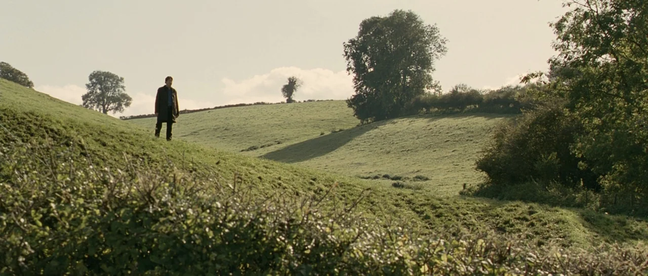 Tamara Drewe (2010) movie still: wide — A rolling green hillside under a bright, cloudy sky with scattered mature trees. A man stands alone on the…
