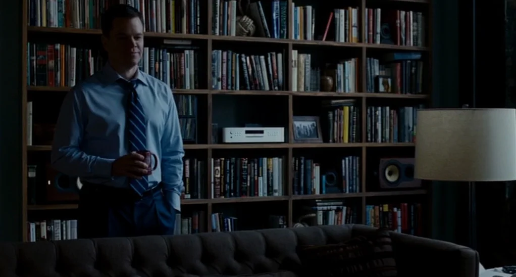 The Adjustment Bureau (2013) movie still: medium — A dark home library features floor-to-ceiling wooden shelves packed with numerous books. A man…
