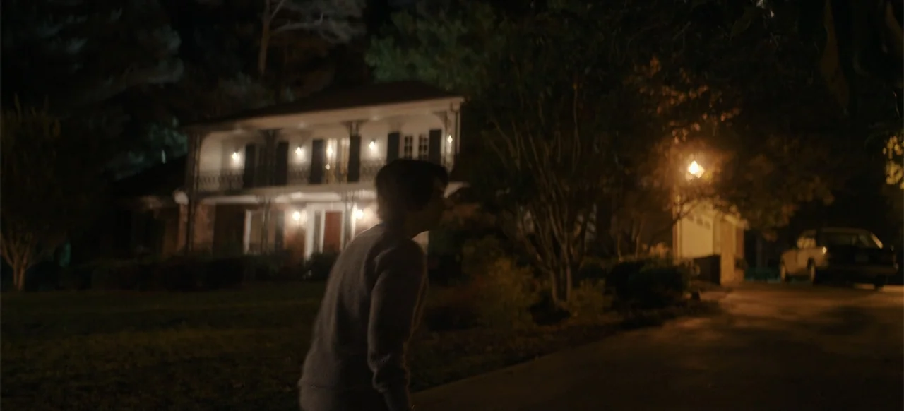 My Best Friend's Exorcism (2022) movie still: medium — A two-story house stands at night behind a dark, grassy front lawn. A person wearing a fuzzy…