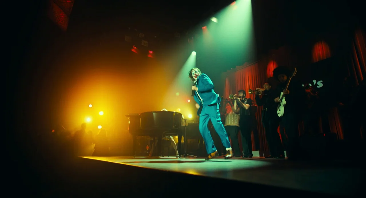 Joker: Folie à Deux (2024) movie still: fullbody — A dimly lit club stage features a man dancing in a blue suit. He performs before a band set…