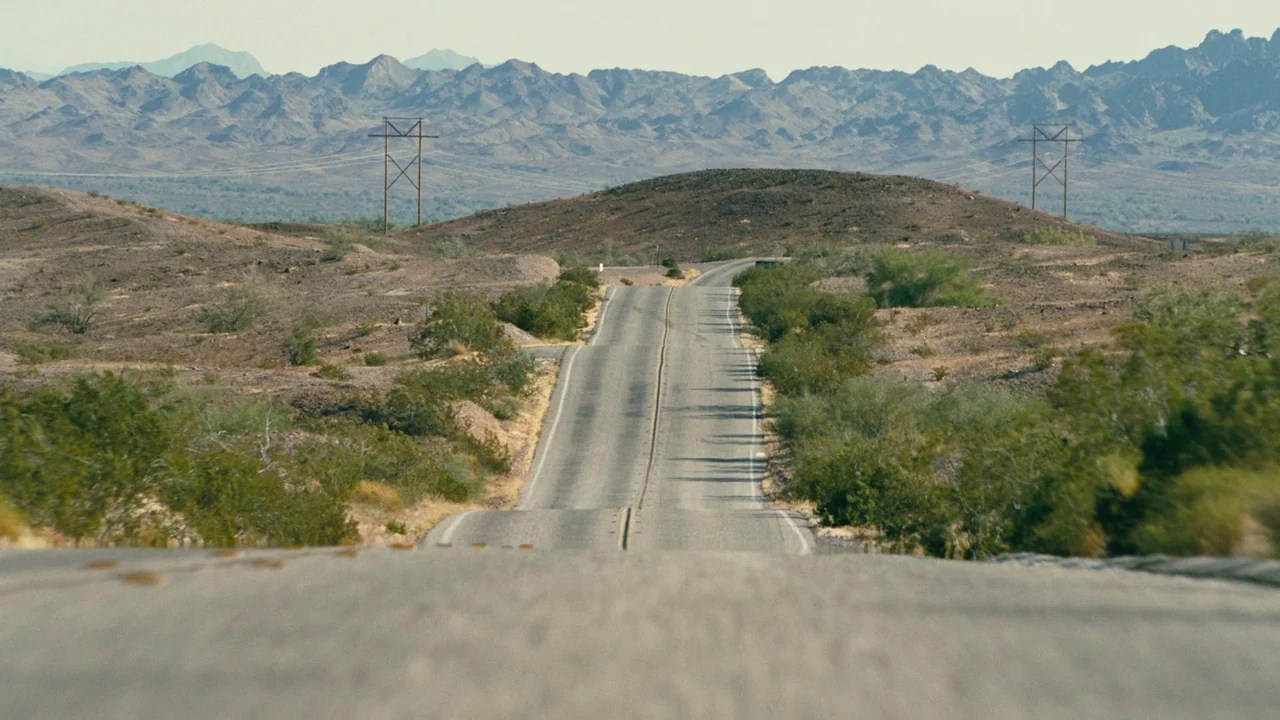One Battle After Another (2025) movie still: establishing — An asphalt road cuts through a vast, arid desert under a bright sky. Power lines and…