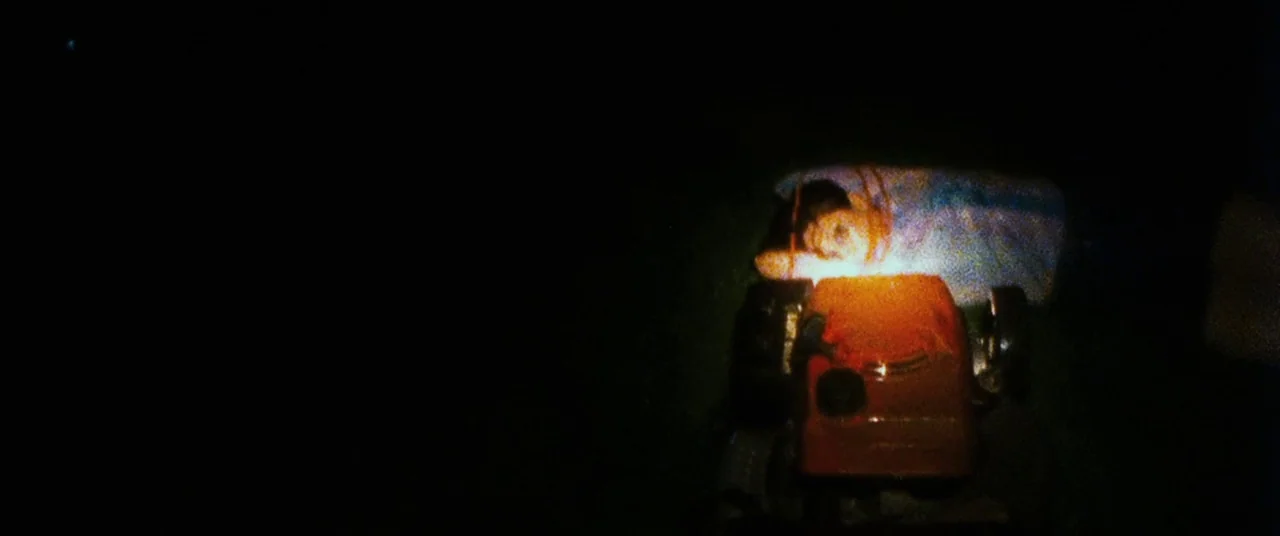 Sinister (2012) movie still: wide — A dark outdoor space is lit by a single point of light at night. A young boy rests on the ground beside a red…