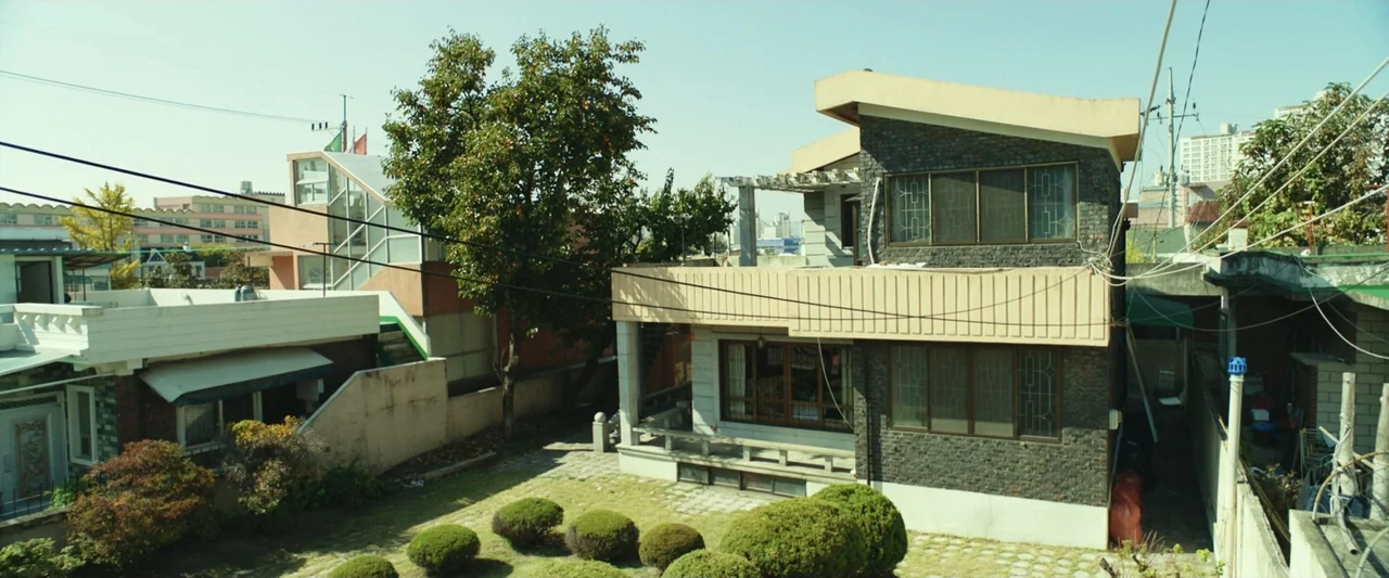 The Gangster, the Cop, the Devil (2019) movie still: establishing — A two-story brick residential house features a flat roof, large windows, and a…