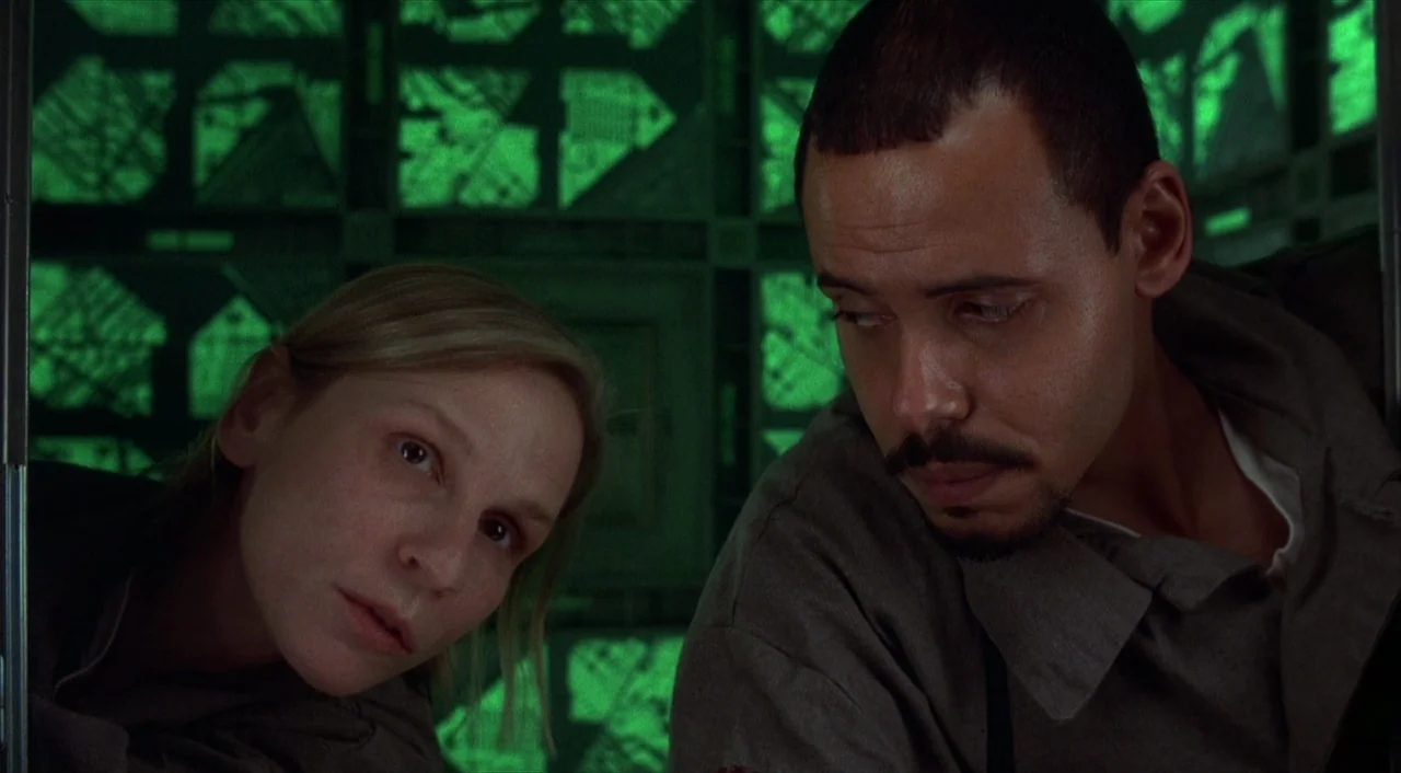 Cube (1997) movie still: medium — A dark enclosure features glowing green walls with an intricate grid pattern. A man and a woman look downward…