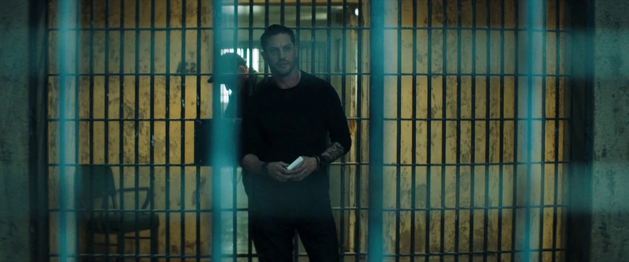 Venom (2018) movie still: medium — An interior space features iron bars dividing the foreground from the main room. A man stands between the bars…