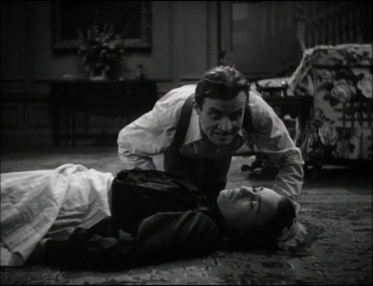 Dracula (1931) movie still: medium — A dimly lit Victorian parlor features ornate furniture and a floral patterned armchair. A man hunches over a…